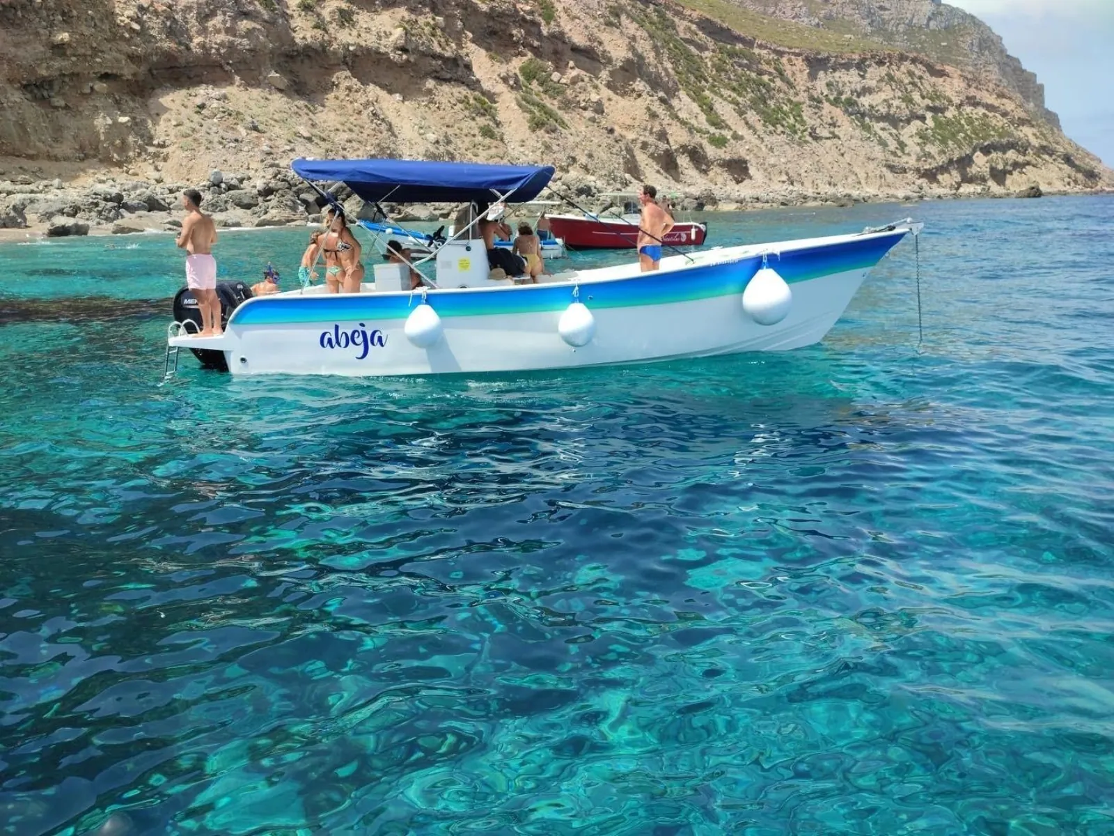 Guests aboard the blue-and-white boat in clear shallow water below the cliffs